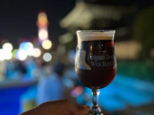 夜のお東さん広場を背景に、ベルギービールのグラスを手に持つ様子