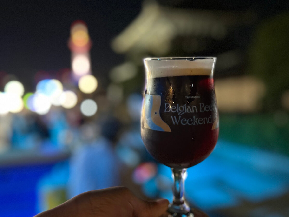 夜のお東さん広場を背景に、ベルギービールのグラスを手に持つ様子